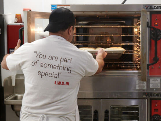 A man in a white t - shirt is putting doughnuts in the oven.