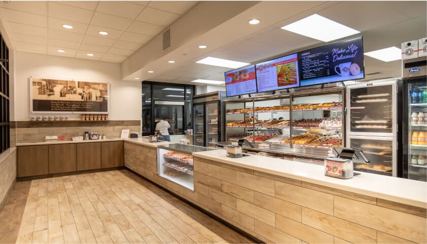 Interior of a modern QSR franchise donut shop with wooden floors, a display case filled with assorted donuts, digital menu screens above, and a refrigerated section containing various drinks.