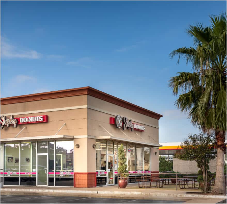 The vibrant donut shop, part of a popular QSR franchise, boasts a welcoming exterior with a catchy sign and large windows. A palm tree sways gently near the inviting outdoor seating area under a clear blue sky.